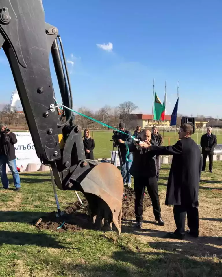 A group of people, including a man in a black coat, participate in a groundbreaking ceremony using a large excavator on a grassy field under a clear blue sky. Several flags are visible in the background, and photographers capture the event.
