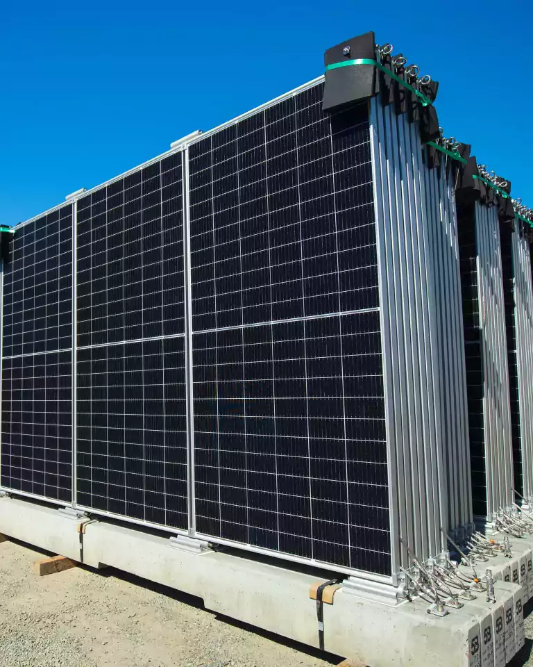 Stacks of large solar panels are arranged outdoors on concrete bases under a clear blue sky, ready for installation at a solar energy facility.
