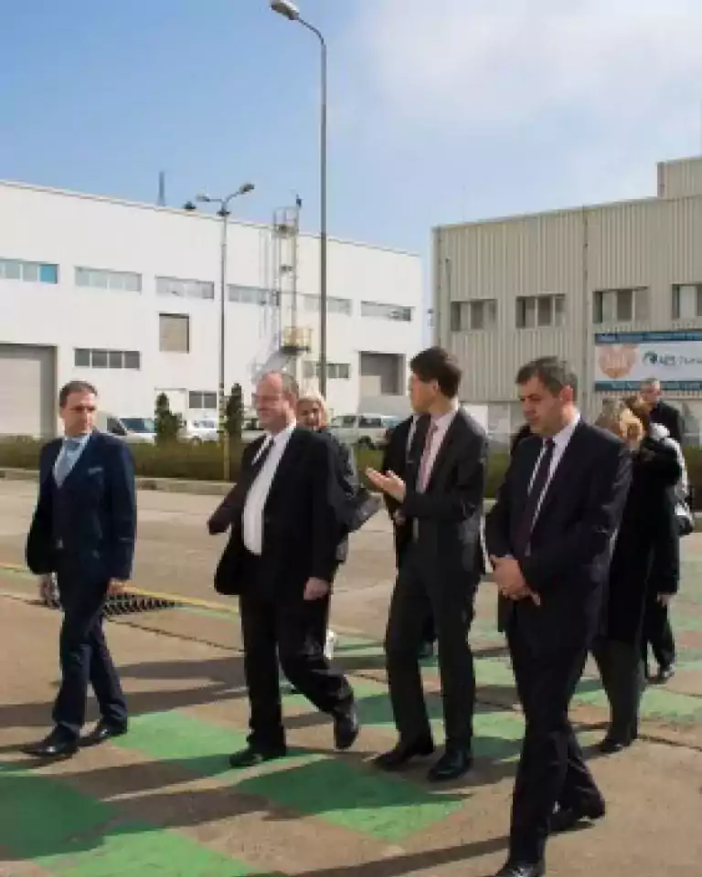 A group of business professionals in formal attire walking and conversing outside an industrial building on a sunny day.
