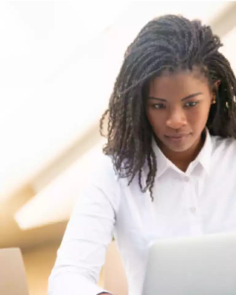 Woman with braided hair wearing a white shirt, focused on working at a laptop in a bright indoor setting.