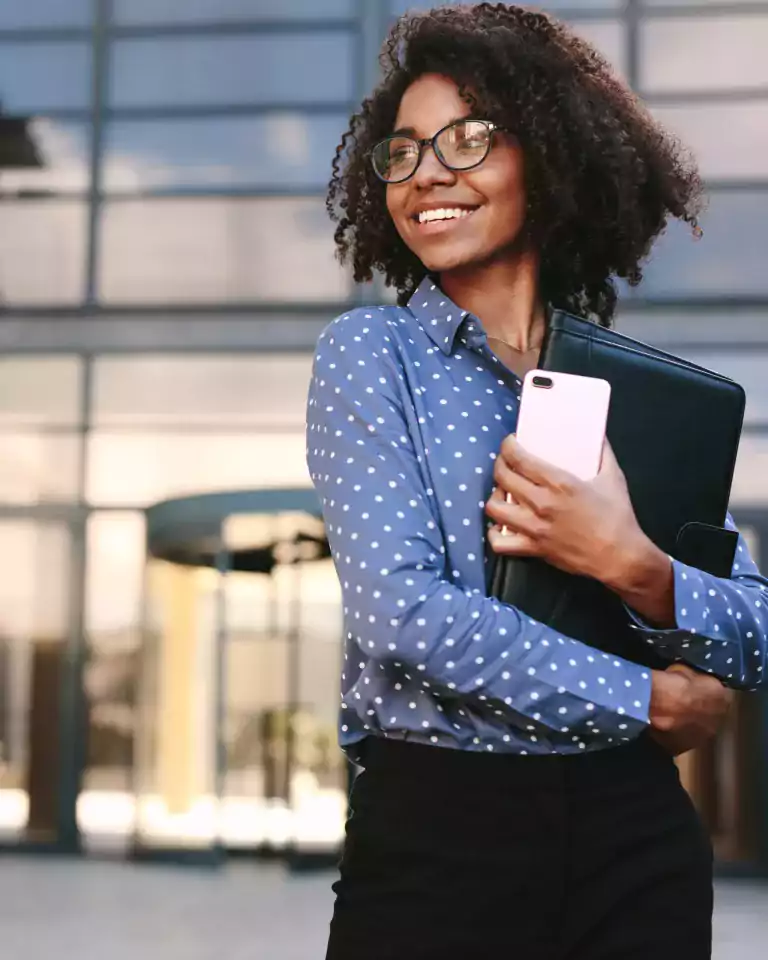 Smiling woman wearing glasses and a blue polka dot shirt stands outside a modern office building, holding a black folder and a pink smartphone.