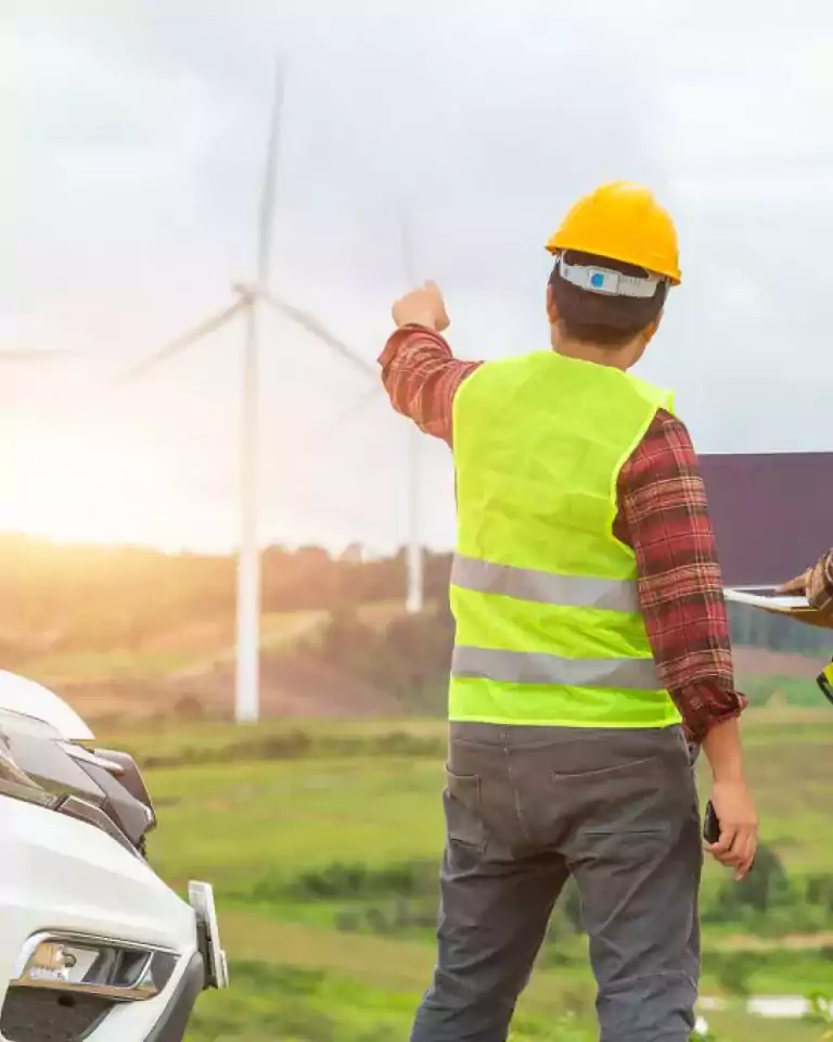 Two engineers wearing yellow safety helmets and reflective vests stand near a white vehicle, examining wind turbines in a green landscape. One engineer points toward the turbines while the other holds a laptop. The sky is cloudy and the sun is setting.