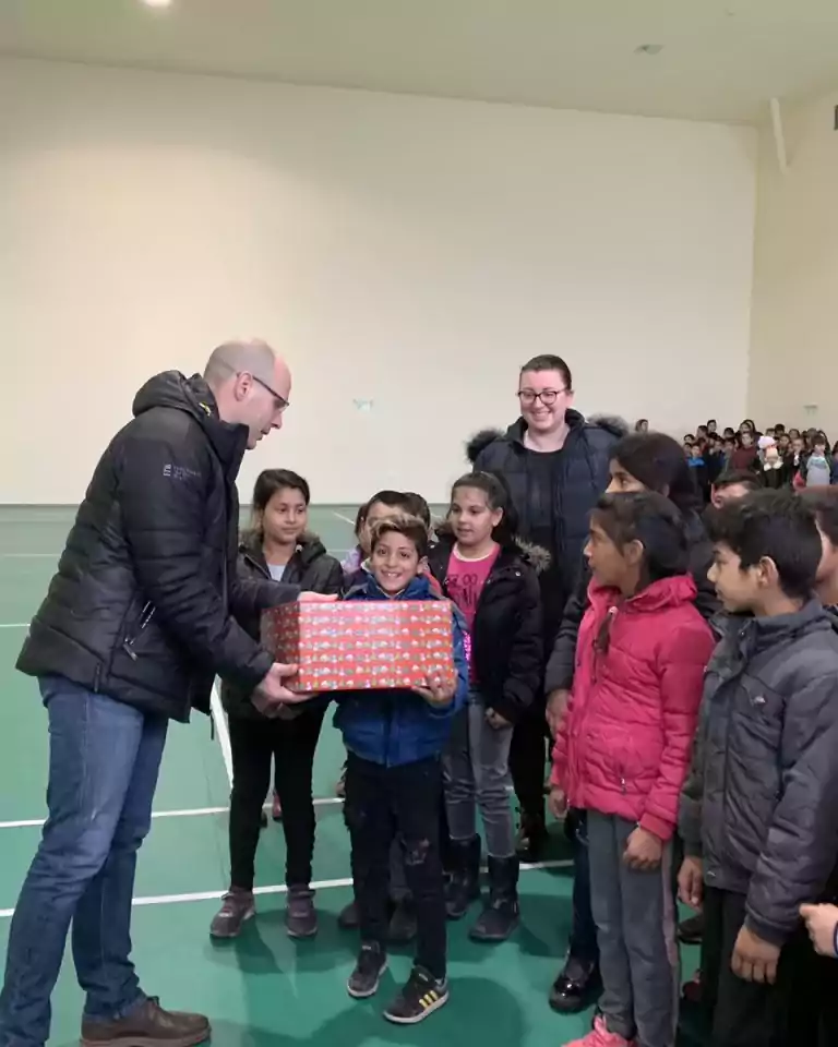 A man in a black jacket hands a gift-wrapped box to a smiling boy standing among a group of children in a gymnasium. Another adult and more children are visible in the background, all wearing winter clothing.