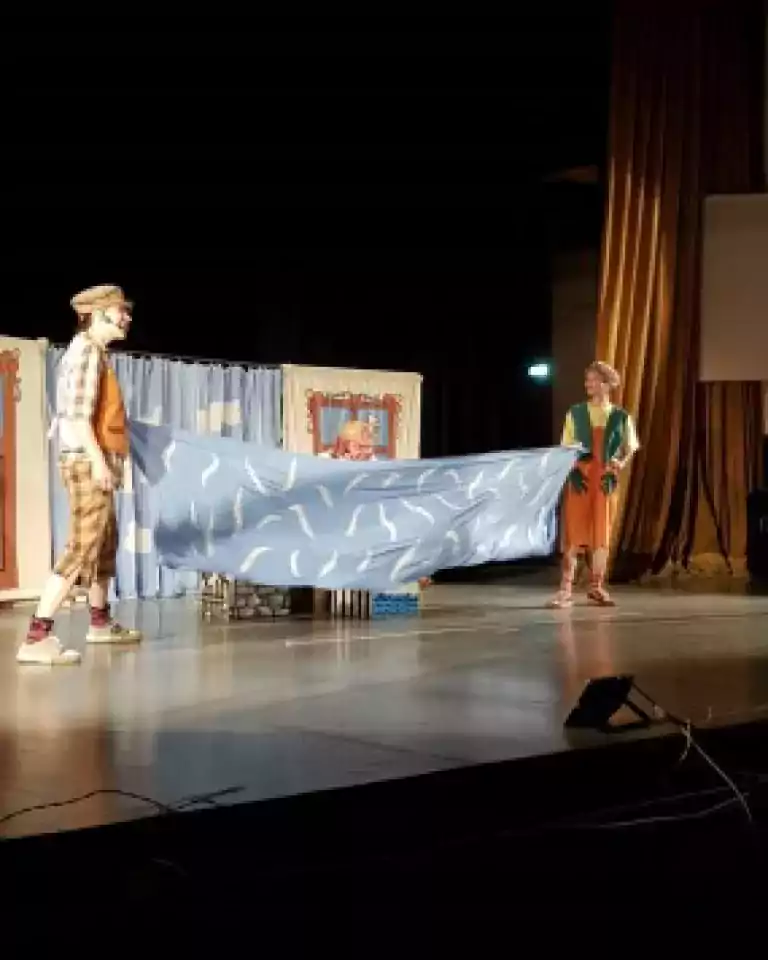 Two actors in colorful costumes hold a blue sheet with white patterns on a theater stage, simulating water. A third actor is partially visible behind the sheet. The background includes stage curtains and set decorations.