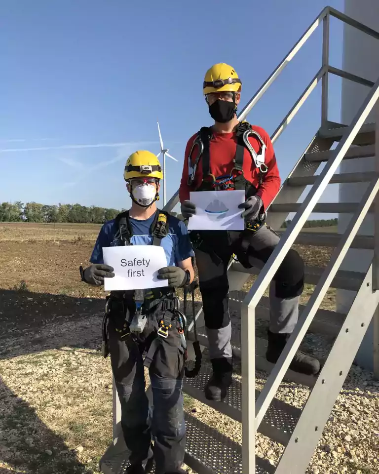 Two workers wearing yellow helmets, safety harnesses, gloves, and face masks stand on metal stairs outdoors near a wind turbine. One holds a sign that says 'Safety first,' emphasizing workplace safety.