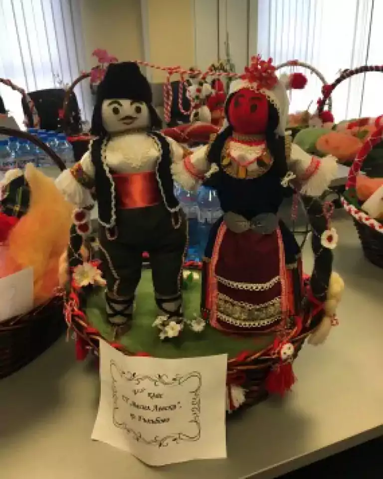 Two handmade dolls dressed in traditional Bulgarian folk costumes are displayed in a decorative basket with festive ribbons and ornaments. The dolls are positioned upright on a table, with a handwritten note in front of the basket.