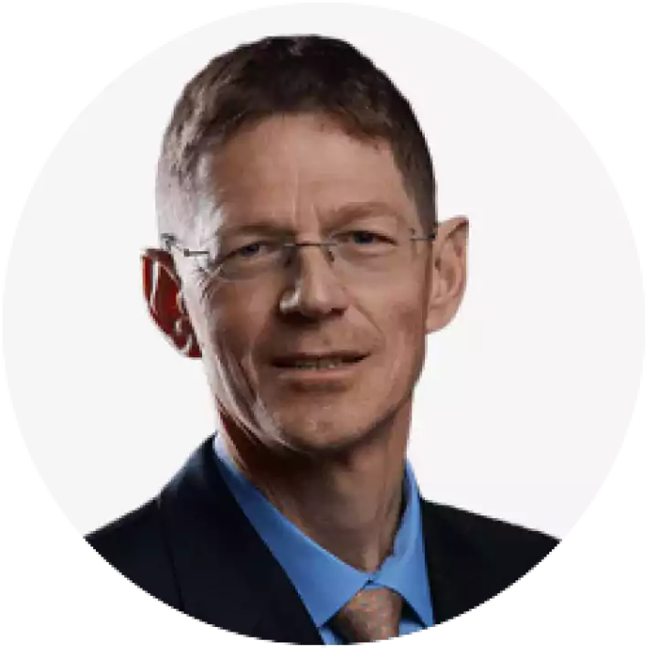 Portrait of a middle-aged man with short brown hair, glasses, wearing a dark suit, blue shirt, and patterned tie, against a plain white background.