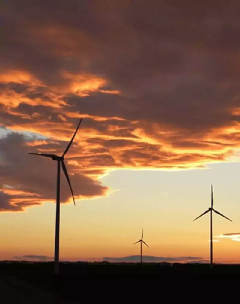 Three wind turbines stand in a field under a dramatic orange and purple sunset sky, illustrating renewable energy in a natural landscape.