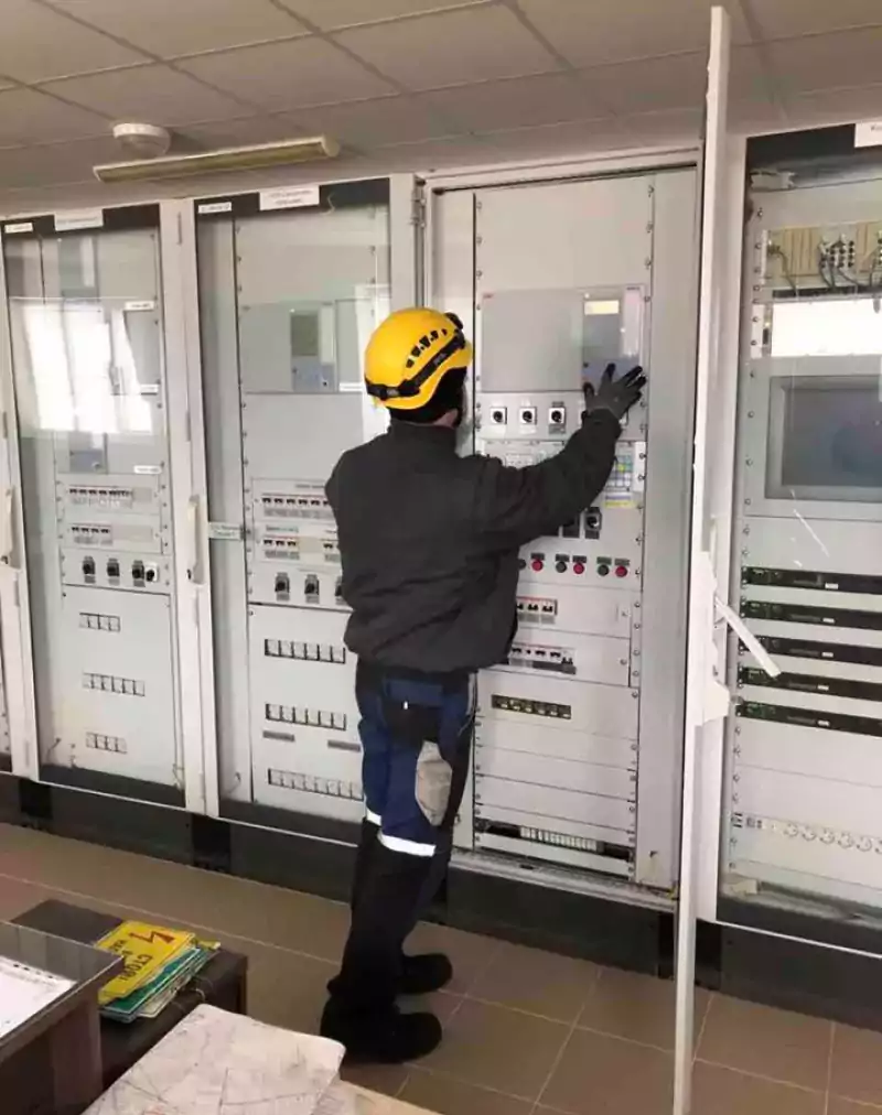 A worker wearing a yellow helmet and protective clothing operates control panels in an industrial control room. The worker is facing the panels, adjusting a switch or button, surrounded by various electrical equipment and instruments.