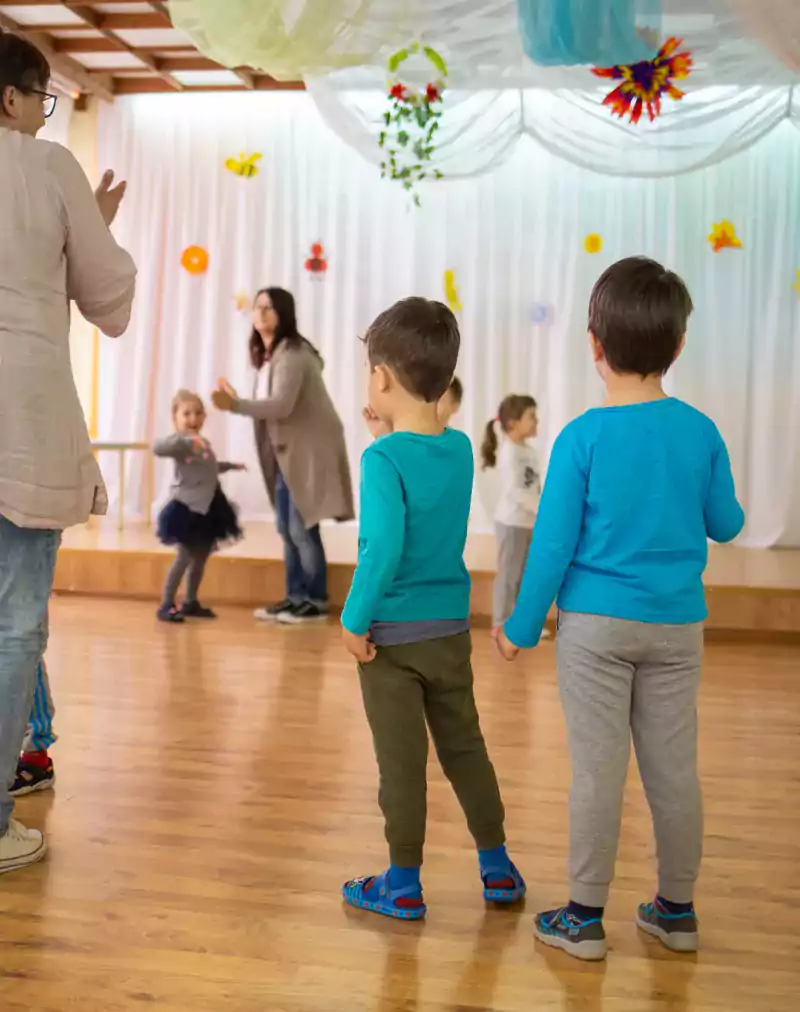 Children and teachers participate in a group activity in a brightly decorated classroom with wooden floors and colorful wall decorations. The children are standing, playing, and interacting in small groups while teachers supervise and guide them.
