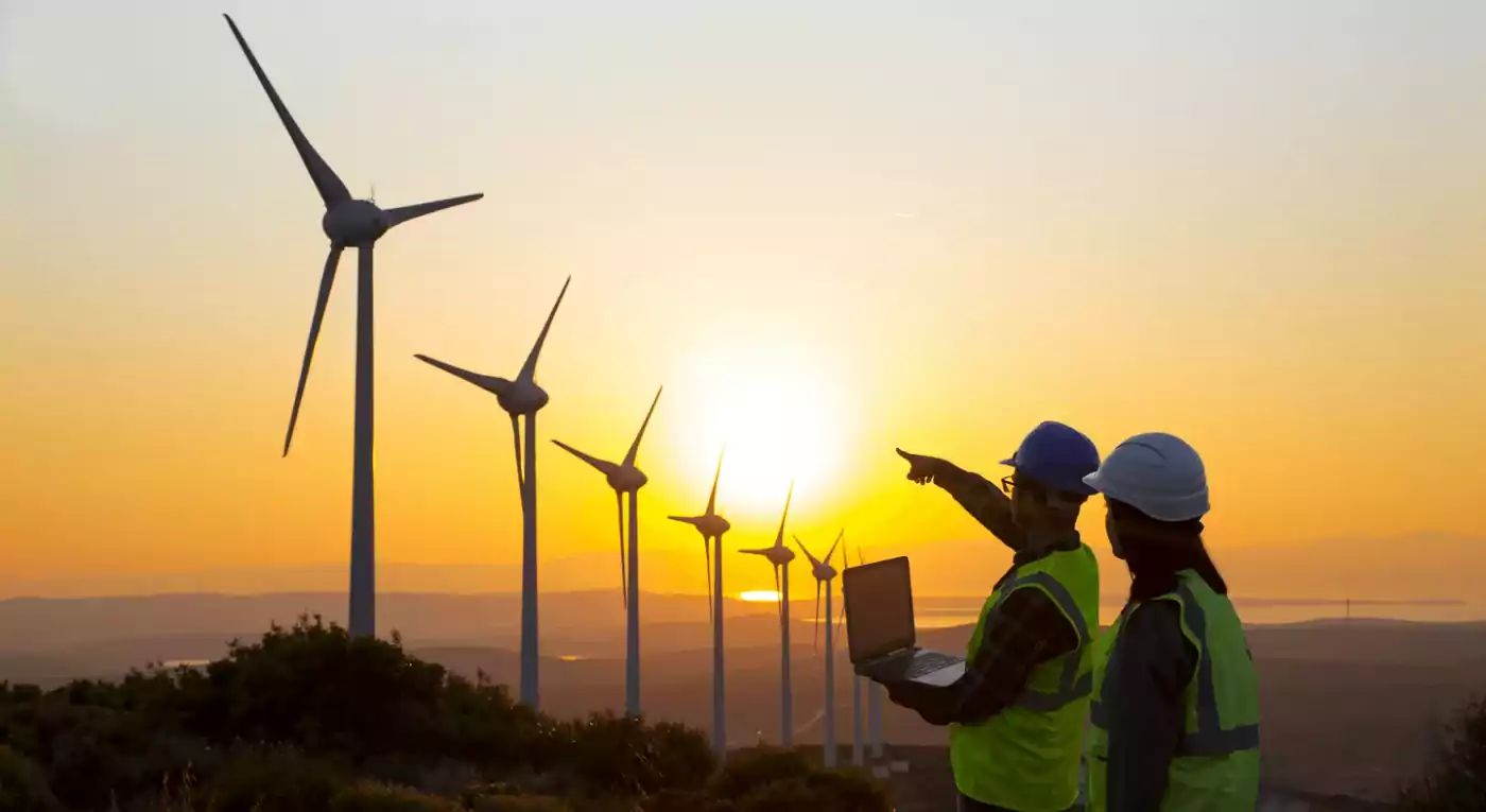 Two engineers wearing safety vests and helmets stand in front of wind turbines at sunset. One engineer holds a laptop while the other points towards the turbines, highlighting renewable energy production in a scenic landscape.