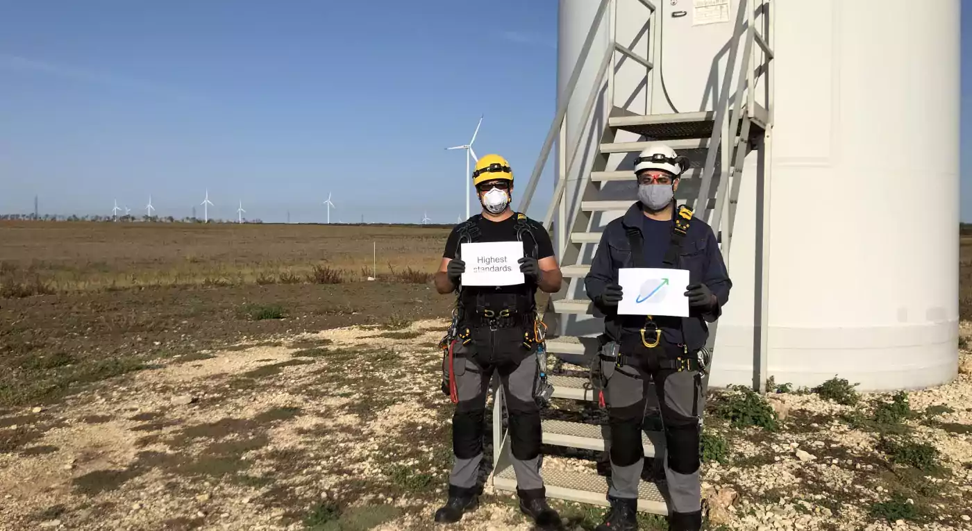 Two wind turbine technicians wearing safety gear and masks stand in front of a wind turbine base, holding signs. The background shows an open field with several wind turbines under a clear blue sky.