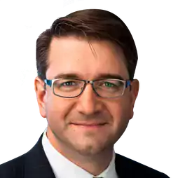 Man with short brown hair wearing glasses, a dark suit, white shirt, and striped tie, smiling at the camera against a transparent background.