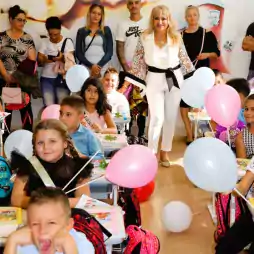 A classroom filled with young children sitting at desks decorated with balloons, books, and backpacks. Some children are smiling, while one boy in the front appears to be yawning or upset. Several adults, including a woman in white, stand at the back, likely teachers and parents, watching the students.