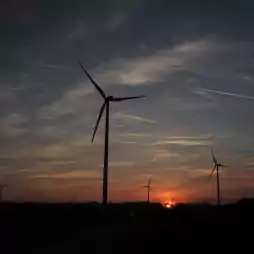 Silhouettes of wind turbines on a field at sunset with a colorful sky and scattered clouds. The sun is low on the horizon, casting an orange glow, and a car is visible on a road in the foreground.