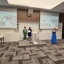 Four people participate in an award ceremony in a modern conference room. Two women stand on a small stage, one holding a certificate and the other a medal. A man in a suit stands on the left, and a woman in a blue dress reads from a paper on the right. Presentation slides are projected behind them.