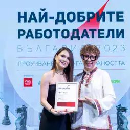 Two women stand on stage holding an award and certificate at a Best Employers Bulgaria 2023 event. The background features a large event banner in Bulgarian and images of chess pieces.