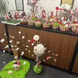 Handmade Martenitsa decorations displayed on a wooden cabinet and green mats, featuring red and white yarn, dolls, flowers, and branches, celebrating the Bulgarian tradition for welcoming spring.