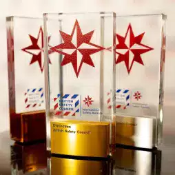 Three British Safety Council International Safety Awards with red star emblems and gold and silver bases, displayed on a reflective surface.