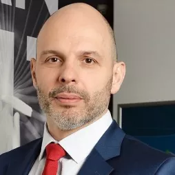 Bald man with a trimmed beard wearing a blue suit, white shirt, and red tie standing indoors in front of a large grayscale sign and computer monitors.