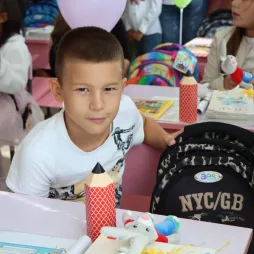 A young boy sits at a classroom desk with a camouflage backpack and school supplies, including a pencil-shaped container and a stuffed toy. Other children are visible in the background, engaged with their own materials in a colorful classroom setting.