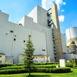 Large industrial power plant building with tall cooling tower, surrounded by green landscaped lawn, trees, and shrubs under a blue sky with clouds.