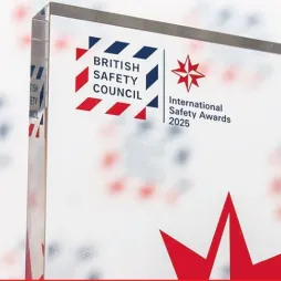 A close-up of a glass award trophy featuring the British Safety Council logo and text reading 'International Safety Awards 2025'. The background is blurred with repeated logos and red star symbols.