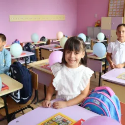 Three young children sit at their desks in a colorful classroom decorated with balloons and school supplies. The students are smiling and appear ready for the school day, with backpacks and books on their desks. The classroom walls are painted pink and have educational posters and cabinets.