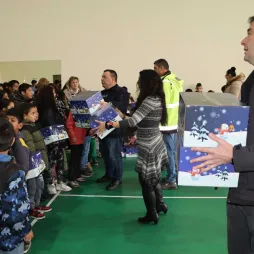 Group of adults distribute festive gift boxes to children standing in line inside a gymnasium. The children are receiving the boxes, which are decorated with winter holiday designs, and appear excited and happy.