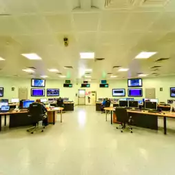 Modern control room with multiple operators seated at desks, monitoring large computer screens and wall-mounted displays. The room is brightly lit with a spacious layout, featuring advanced technology and ergonomic chairs.