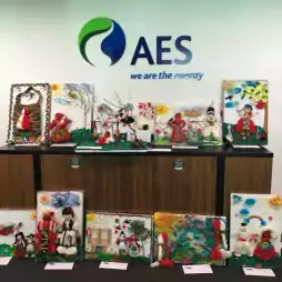 Display of colorful handmade crafts and artwork featuring traditional Bulgarian Martenitsa dolls and spring themes, arranged on a counter and floor in front of an AES logo and the slogan 'we are the energy' on the wall.
