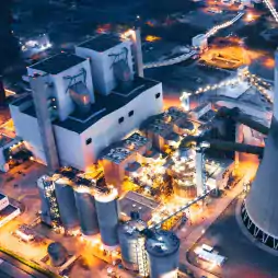 Aerial view of a large industrial power plant at night, illuminated by bright lights, showing multiple cylindrical storage tanks, interconnected pipes, and a main building surrounded by infrastructure.