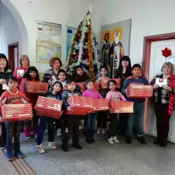 A group of children and adults stand indoors in front of a decorated Christmas tree, holding red gift boxes and envelopes. The setting appears to be a school or community center, with posters and artwork on the walls. The atmosphere is festive and joyful.