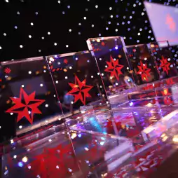 Several clear glass awards with red star designs are displayed on a reflective table under a backdrop of twinkling lights at an indoor event, with a large screen visible in the background.