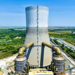 Large cooling tower of a power plant with industrial pipes and structures, surrounded by green fields and a clear blue sky in the background.