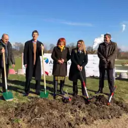 Six people standing outdoors on a grassy field, holding shovels and posing for a photo at a groundbreaking ceremony under a clear blue sky.