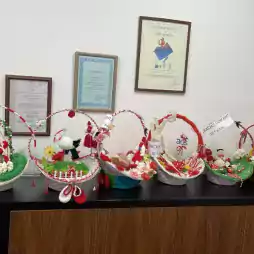 A row of decorative baskets on a black cabinet, each filled with traditional Bulgarian Martenitsa ornaments in red and white, flowers, and festive figures. Framed certificates and awards hang on the white wall above the display.