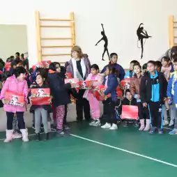 A group of children and a few adults stand together in a gymnasium, holding red gift boxes with festive designs. Some children are smiling and talking, while others look at the camera. The gym has green flooring and wall bars, with silhouettes of dancers on the wall.