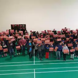 Large group of children and adults standing in a gymnasium, holding red and white gift boxes, facing the camera and smiling. The gym has a green floor with white lines and a plain beige wall in the background.