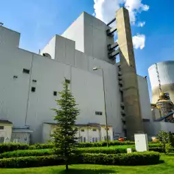 Modern power plant with large industrial buildings, a tall chimney, and a cooling tower, surrounded by green landscaped lawns and small trees under a blue sky with scattered clouds.