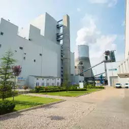 Modern industrial power plant with large white buildings, a cooling tower emitting steam, landscaped green lawns, and parked vehicles under a blue sky.