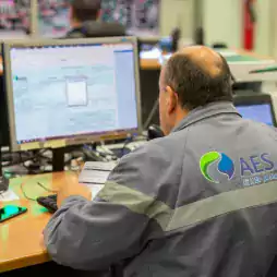 A worker wearing a gray AES uniform sits at a desk and operates a computer in a control room, with multiple monitors and control panels visible in the background.