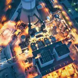 Aerial view of an illuminated industrial power plant at night, showing large cooling towers, storage tanks, and various industrial buildings with bright lights and surrounding roads.