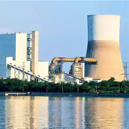 A large power plant with a cooling tower and industrial buildings stands beside a body of water, with green trees in the foreground and clear blue sky in the background.