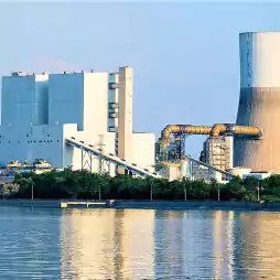 A large industrial power plant with a cooling tower and several buildings is situated near a body of water, surrounded by greenery under a clear blue sky.
