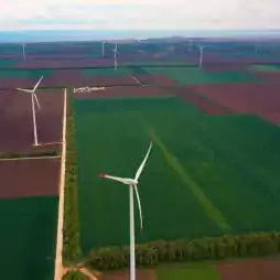 Aerial view of a wind farm with multiple wind turbines spread across large green and brown agricultural fields near the coastline under a cloudy sky.