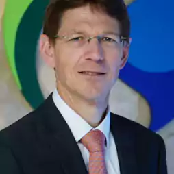 Man wearing a dark suit, white shirt, and orange tie, with glasses and short brown hair, standing in front of a blue and green abstract background.