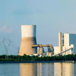 A large power plant with a cooling tower, industrial buildings, and power lines is situated near a body of water under a clear sky.