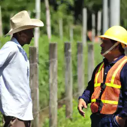 A farmer wearing a straw hat and a construction worker in a yellow helmet and orange safety vest are having a conversation outdoors near a wire fence with green vegetation in the background.