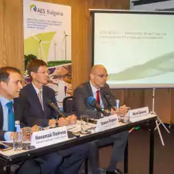 Three men in formal attire sit at a conference table with microphones and nameplates, participating in an AES Bulgaria energy event. A presentation screen and promotional poster with wind turbines are visible in the background.